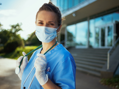 Portrait of a nurse standing in front of a hospital in mask and gloves wearing a stethoscope in 