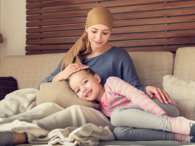 Young female cancer patient in headwrap sitting on a couch, looking down at her daughter who is laying in her lap.