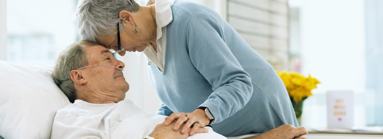 A wife stands next to her husband's hospital bed, touching her forehead to his