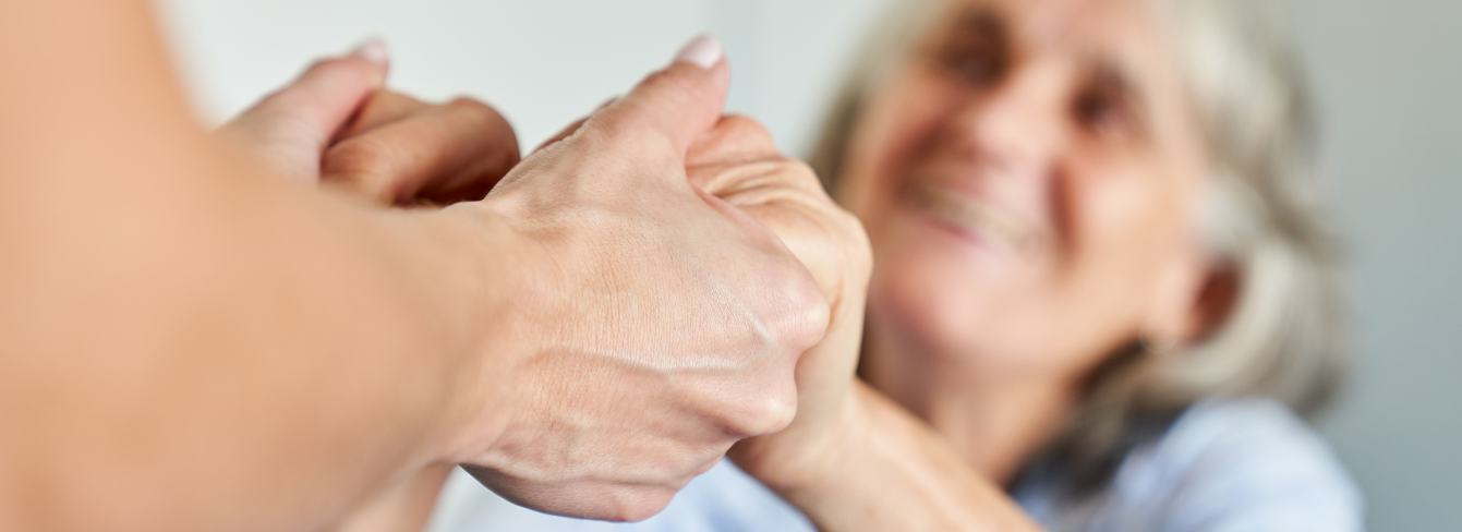 A caregiver holds hands with a patient, whose face is visible in the background A caregiver holds hands with a patient, whose face is visible in the background