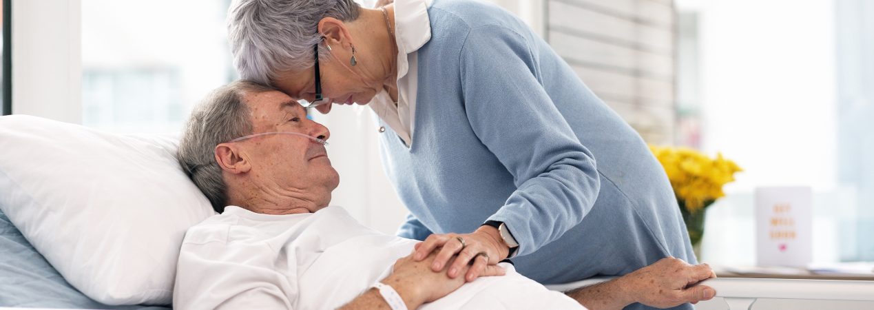 A wife touches her forehead to her husband's, who is in a hospital bed. A wife touches her forehead to her husband's, who is in a hospital bed.