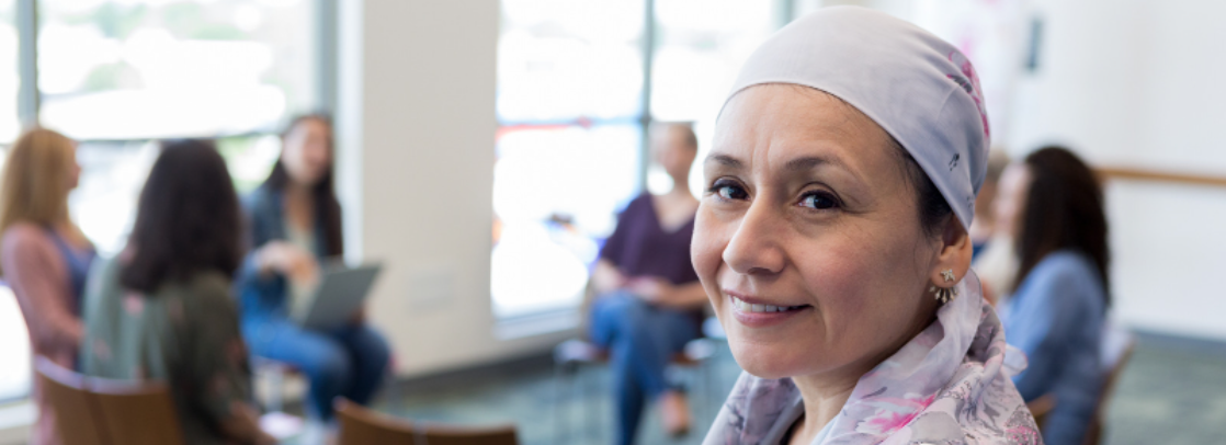 Smiling female cancer patient in headwrap standing in a room with a support group gathered in the background Smiling female cancer patient in headwrap standing in a room with a support group gathered in the background