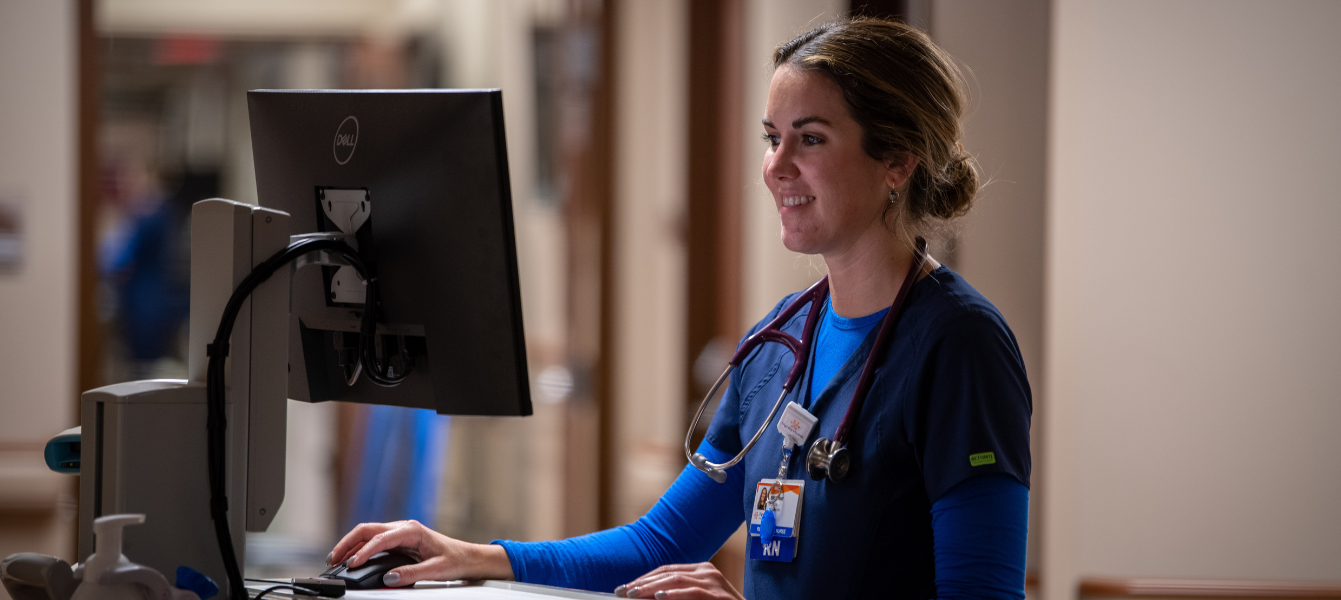 Smiling female nurse working at computer. Young, female French Smiling female nurse working at computer. nurse in mask standing in a patient room