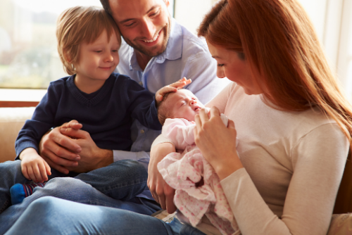Young smiling mother sitting with her husband and son on the couch while holding their newborn baby