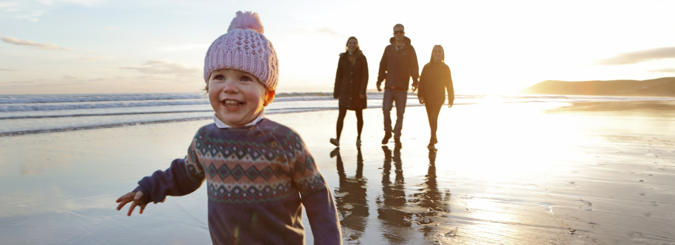 A smiling toddler walks on the beach with their family behind them